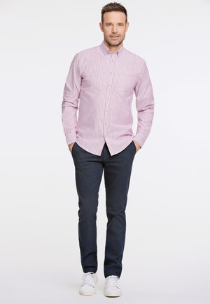 Camicia a righe rosa chiaro e bianca con bottoni, pantaloni blu navy, sneakers bianche. Modello in piedi con le mani nelle tasche contro uno sfondo semplice.