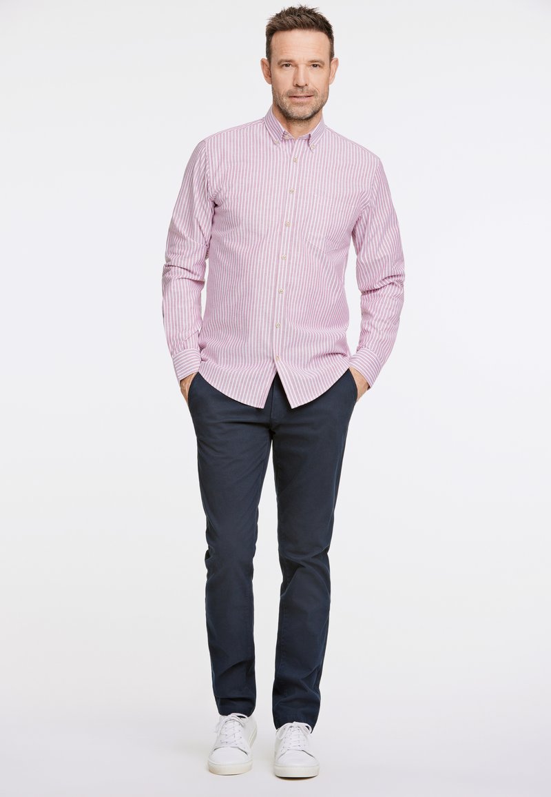 Camisa de botones rayada en rosa claro y blanco, pantalones azul marino, zapatillas blancas. El modelo está de pie con las manos en los bolsillos frente a un fondo liso.