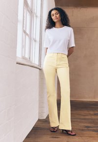 Jean jaune évasé avec poches avant, assorti à un haut blanc à manches courtes. Le mannequin se tient près d'une fenêtre sur un sol en bois.