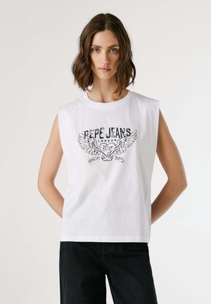 Donna con capelli castani mossi, che indossa un top bianco senza maniche con una grafica nera "Pepe Jeans London" e pantaloni neri, in piedi con le mani dietro la schiena.