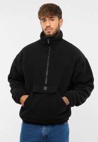 Čierny fleecový pulóver s vysokým golierom, polovičným zipsom vpredu, náprsné vrecko a textúrovaná tkanina. Má rebrované manžety a voľný strih.