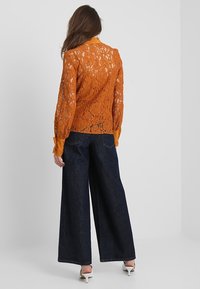 Top in pizzo arancione con maniche lunghe, motivo floreale dettagliato e colletto a tinta unita. Abbinato a pantaloni in denim scuri e larghi.