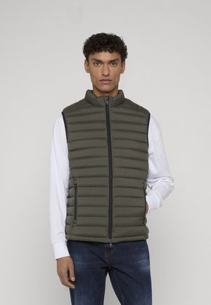 Mężczyzna ubrany w oliwkowozieloną pikowaną kamizelkę bez rękawów na białej koszuli z długim rękawem i niebieskich dżinsach, stojący na tle jednolitego jasnego tła.