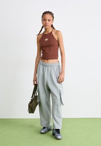 Brun halterneck crop-top med logo, grå cargo-sweatpants og grå sneakers. Holder en grønn veske. Minimalistisk design med synlige teksturer.