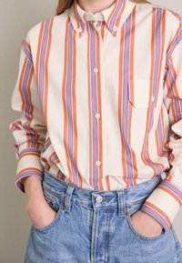 Randig skjorta i kräm, orange och blå; har en ficka, manschetter och en avslappnad passform, tillsammans med högmidjade denimshorts.