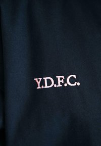 Jūrų jėgos audinys, su išsiuvinėtomis raidėmis "Y.D.F.C." šviesiai rožine spalva. Tekstūra lygi, su įstrižai raštu.