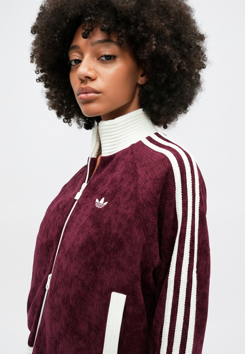 Bordeaux cordjacket met een hoge geribbelde kraag, witte rits en drie witte strepen op de mouwen. Bevat een wit Adidas-logo op de borst.