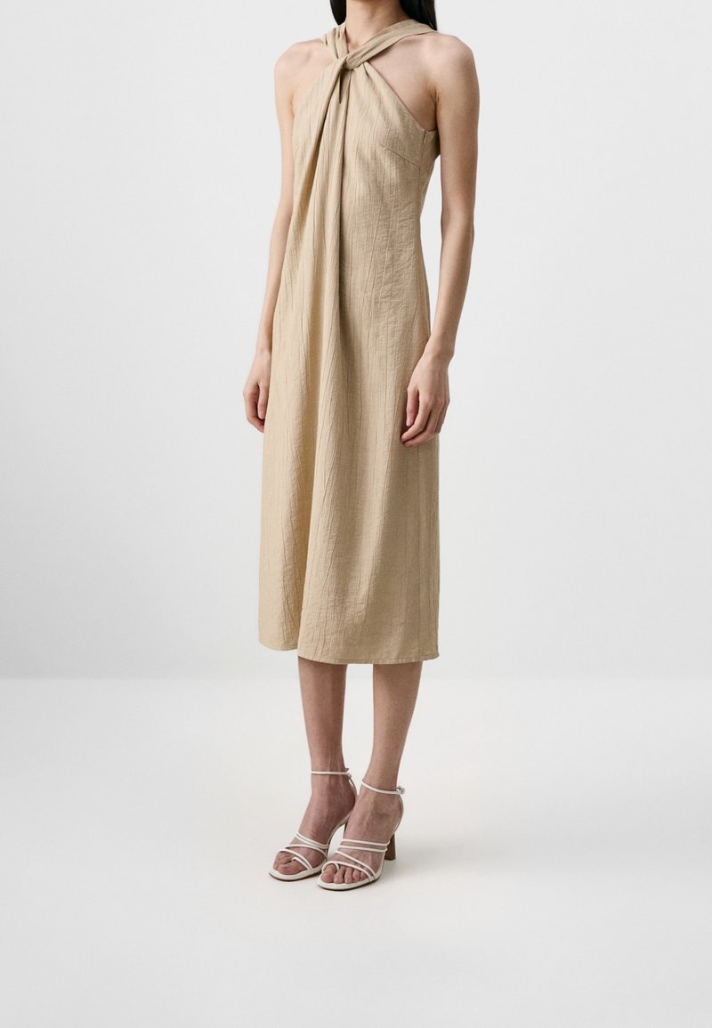 Femme portant une robe midi beige sans manches avec un col halter torsadé et des sandales blanches à talons avec brides, debout devant un fond uni.