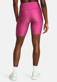 Mujer vestida con pantalones cortos deportivos ajustados de color rosa brillante, calcetines blancos y zapatillas blancas, de pie con la espalda hacia la cámara sobre un fondo sencillo.