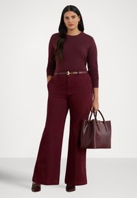 Γυναίκα που στέκεται φορώντας πουλόβερ μπορντό, παντελόνι με φαρδιά μπατζάκια, ταιριαστή ζώνη και τακούνια, κρατώντας μια τσάντα μπορντό με υφή.