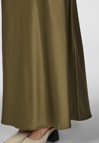 Robe maxi en satin vert olive avec une texture lisse et un ourlet incurvé, accompagnée de chaussures à plateforme beiges à bout ouvert.