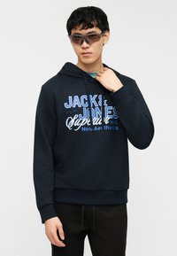 Νέος άνδρας που φοράει σκούρα μπλε μπλούζα με κουκούλα και το λογότυπο "Jack & Jones" και μαύρο παντελόνι, διορθώνοντας το γιακά της κουκούλας, σε απλό λευκό φόντο.