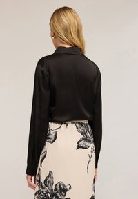 Femeie blondă întoarsă, purtând o bluză lungă neagră cu mâneci și o fustă crem cu imprimeu floral mare negru, pe un fundal simplu.
