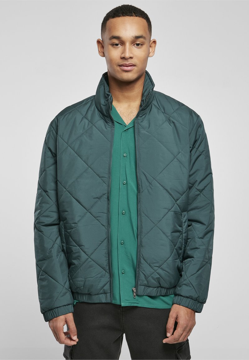 Jeune homme portant une veste matelassée vert foncé sur une chemise verte à boutons, debout devant un fond uni gris clair.