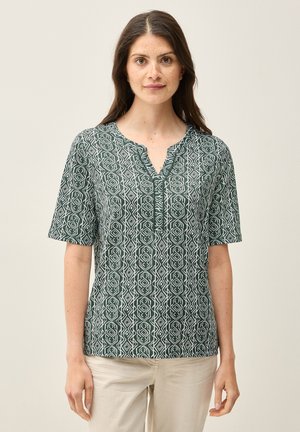 Blouse verte à motifs avec des manches courtes, dotée d'un col en V et d'un design géométrique blanc, fabriquée dans un tissu léger.