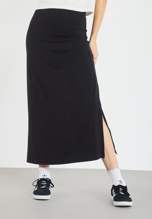 Personne portant une jupe midi noire avec une fente latérale, des chaussettes blanches, des baskets noires et un haut blanc, debout devant un fond uni.