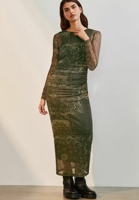 Olīvzaļš maksi kleita ar garām caurspīdīgām piedurknēm, krokotām detaļām un paisley rakstiem. Saskaņota ar melniem masīviem potīšu zābakiem.