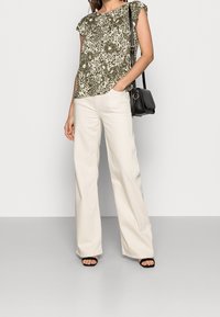 Mujer vestida con una blusa floral verde oliva, pantalones anchos crema, sandalias negras y llevando un bolso negro al hombro, de pie con una mano en el bolsillo.