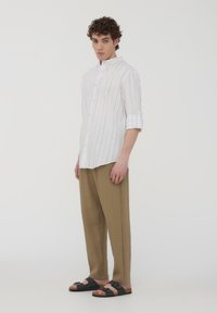 Camicia bianca a righe con colletto alla coreana, abbinata a pantaloni larghi color tabacco e sandali neri, in piedi davanti a uno sfondo neutro.