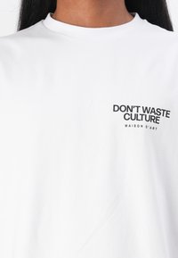 Weißes Baumwoll-T-Shirt mit schwarzem Text, der "DON'T WASTE CULTURE" und "MAISON D'ART" auf der linken Seite zeigt. Einfaches Design und lockere Passform.