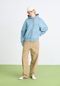 Felpa con cappuccio azzurro chiaro con tasca frontale, abbinata a pantaloni larghi beige. Sneakers bianche con strisce nere. Sfondo uniforme.