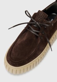 Chaussure en daim marron avec bout arrondi, lacets marron assortis et semelle en caoutchouc texturée de couleur beige clair. Présente un logo en relief sur la languette.
