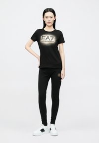Schwarzes Baumwoll-T-Shirt und Leggings-Set mit metallischem Logo-Design. Das Modell trägt weiße Turnschuhe mit schwarzen Akzenten.