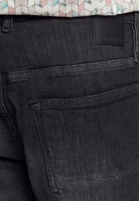 Schwarze Jeans aus Denim mit glatter Textur, ausgestattet mit zwei Gesäßtaschen und einem schwarzen Lederetikett am Bund.