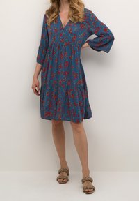 Sininen kukkamekko V-kaula-aukolla, kolmen neljänneksen hihoilla, kerroksellisella muotoilulla ja valuva siluetti. Yhdistetty ruskeisiin punottuihin sandaaleihin.