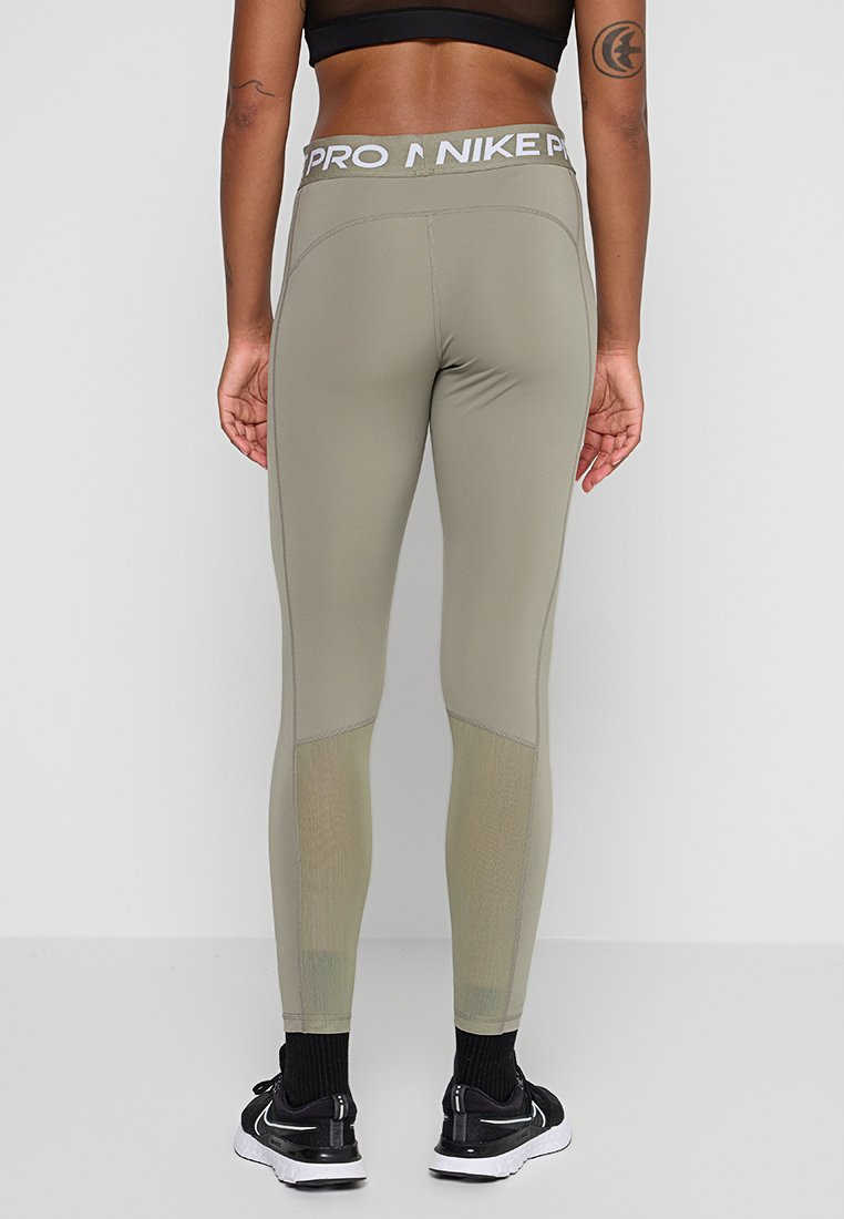 Mujer de pie de espaldas, vistiendo leggings Nike Pro en un tono verde oliva claro con paneles de malla, sujetador deportivo negro, calcetines negros y zapatillas negras.