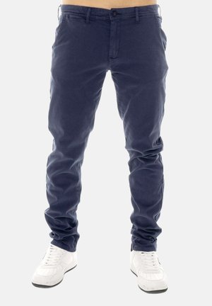 Uomo che indossa pantaloni slim fit blu navy e sneakers bianche, in piedi davanti a uno sfondo chiaro uniforme, ritagliato in vita.