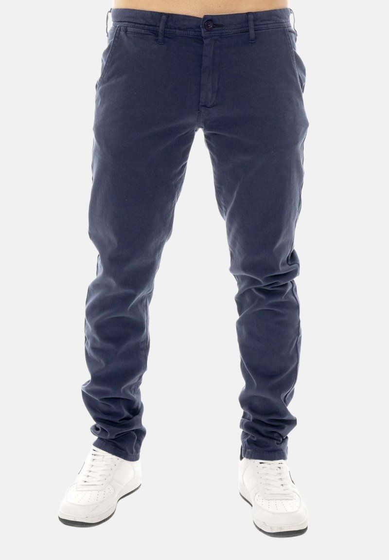 Homme portant un pantalon slim bleu marine et des baskets blanches, debout devant un fond clair uni, cadré à la taille.