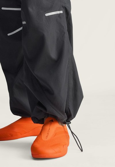 Crne široke hlače s podesivim vezicama na gležnjevima, nošene preko sjajnih narančastih slip-on cipela s "Stella McCartney" brandiranjem.