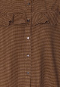 Brun rutigt blus med en volangdetalj vid axlarna, som har svarta knappar framtill och textilmaterial med struktur.