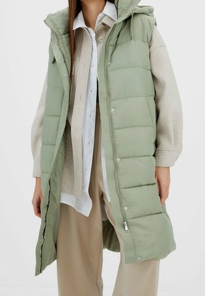 Persoon die een lange lichtgroene gewatteerde capuchonvest draagt over een beige cardigan, wit overhemd en beige broek met brede pijpen.