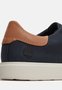 Canvas sneakers in donkerblauw met een tan leren accent op de hiel. Witte rubberen zool; heeft een geborduurd logo aan de zijkant.