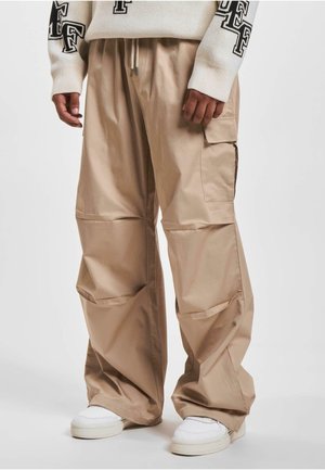Pantalons cargo beiges en tissu léger, avec une coupe décontractée, de grandes poches latérales et une taille élastique. Portés avec des baskets blanches.
