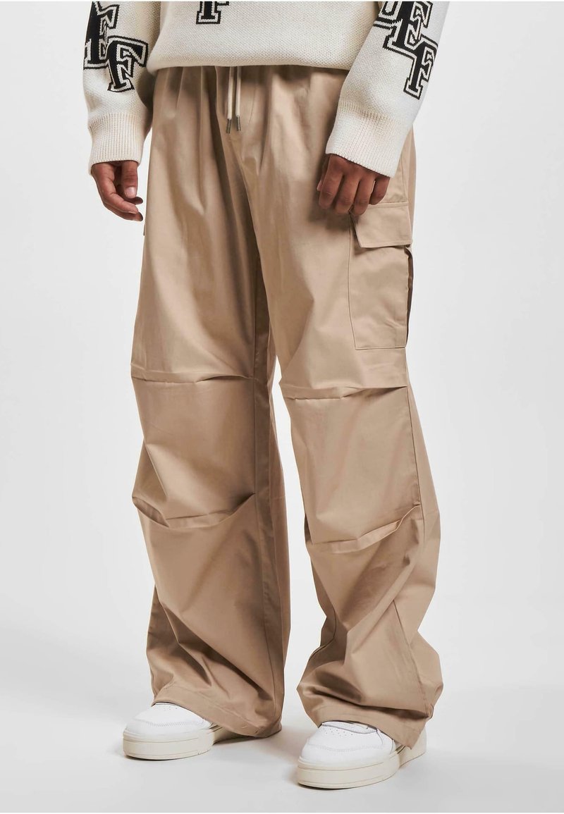 Pantaloni cargo bej dintr-un material ușor, cu un croi lejer, buzunare laterale mari și talie elastică. Purtați cu adidași albi.