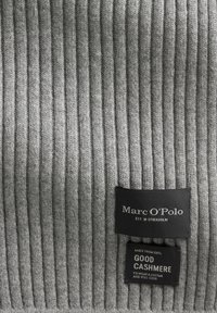 Tissu en cachemire gris côtelé avec deux étiquettes : une noire avec "Marc O'Polo" et une autre indiquant "100 % CACHEMIRE DE QUALITÉ". Texture douce.