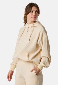 Felpa beige a coste con tasca frontale, cappuccio e cordino. Texture morbida e vestibilità ampia. Abbinata a pantaloni a coste coordinati.