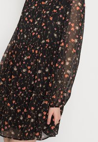 Vestido preto com flores, com mangas longas e transparentes. Apresenta um design em camadas, flores laranjas e brancas espalhadas, e detalhe de cintura elástica.