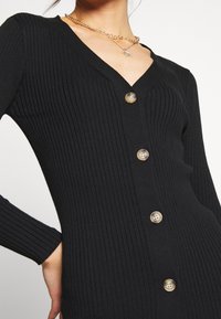 Schwarze gerippte Strickjacke mit V-Ausschnitt und fünf beigen Knöpfen auf der Vorderseite. Verfügt über lange Ärmel und eine taillierte Passform. Strukturierter Stoff.