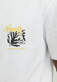 Bijela majica s crnim uzorkom lišća i žutim natpisima "Ninety" i "Jack & Jones ©1990" s lijeve strane. Mekana tkanina.