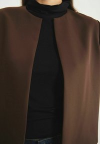 Veste extérieure légère marron avec un design minimaliste, à l'avant ouvert et à la texture lisse. Portée sur un haut noir à col montant.