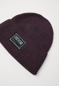 Pletená čiapka v.hlbokom purpurovom odtieni, s rebrovanou textúrou a výraznou obdĺžnikovou etiketou s logom "Versace Jeans Couture" v čiernej a bielej farbe.