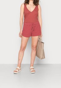 Top rojo sin mangas con escote en V, combinado con pantalones cortos rojos con cordón. Modelo lleva sandalias de plataforma beige y sostiene un bolso tipo tote beige.