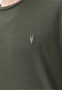 Oliwkowy t-shirt z okrągłym dekoltem i krótkimi rękawami. Posiada małą białą grafikę czaszki wyszytą na lewej piersi.