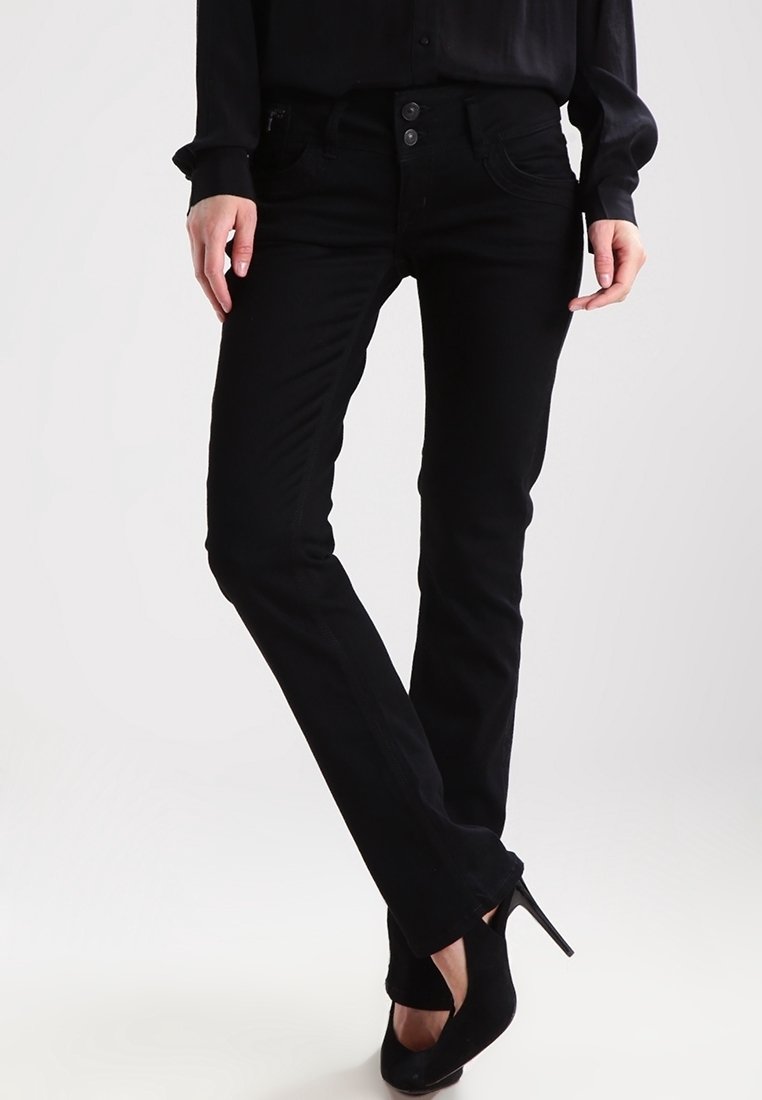 Femme debout, jambes croisées, portant un jean noir ajusté, un chemisier noir et des chaussures à talons noirs sur fond uni.