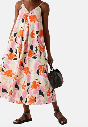 Femme portant une robe sans manches à imprimé floral avec des accents roses, orange et noirs, tenant un sac à main noir et portant des sandales noires.