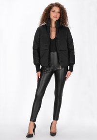 Fekete puffdzseki magas gallérral, steppelt mintával. Fényes fekete leggingsekkel és szegecses magassarkú cipővel kombinálva, fehér háttér előtt.